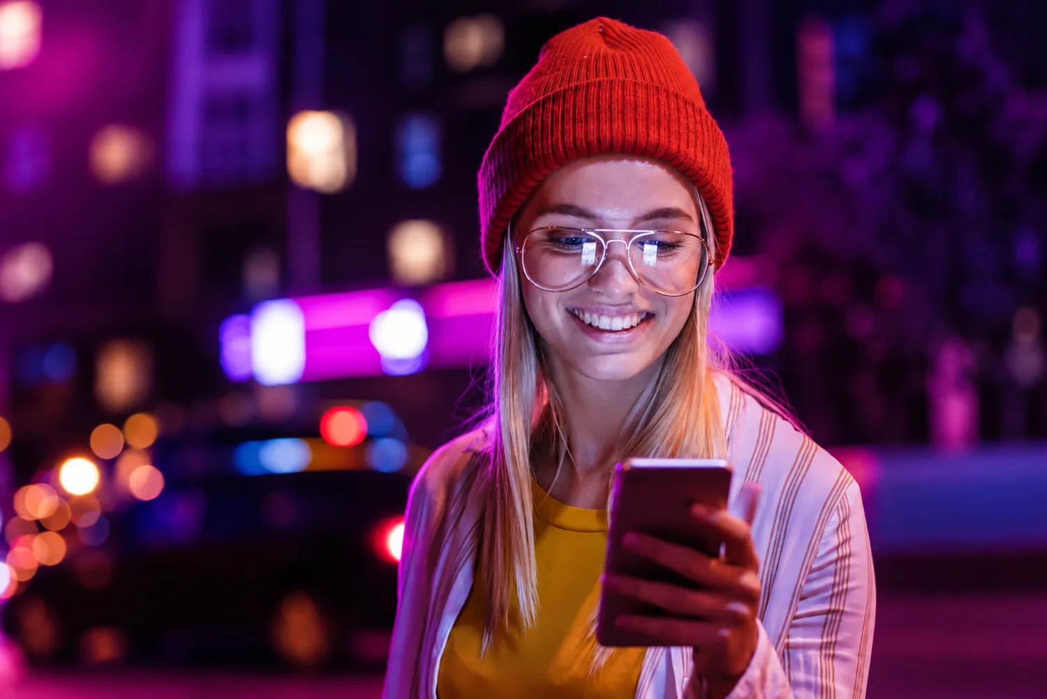 Lachende junge Frau mit roter Mütze schaut nachts auf ihr Smartphone